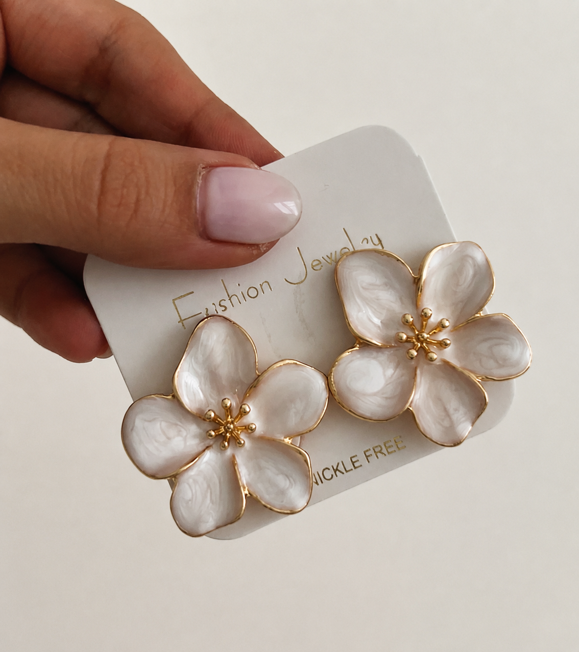 White enamel flower stud earrings with gold trim, held in hand on "Fashion Jewelry" branded card
